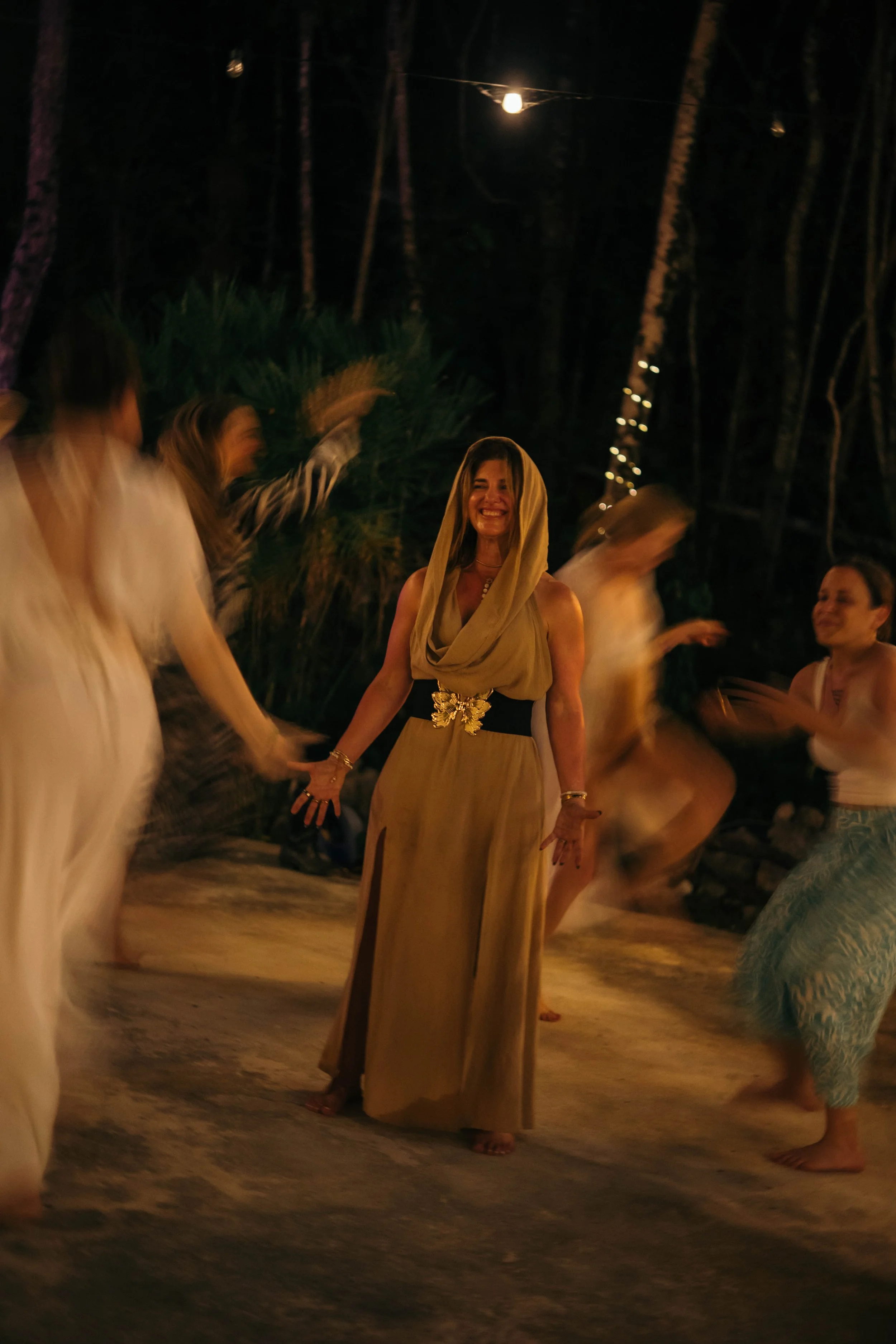 People dancing at an outdoor night event, with a woman in a beige dress and headscarf smiling in the center, surrounded by blurred dancers.