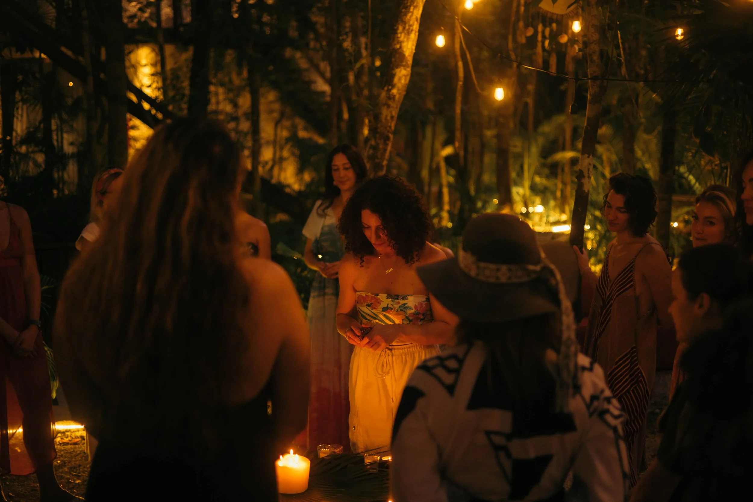 Group of women gathered outdoors at night, illuminated by hanging string lights and candles, with lush trees in the background.