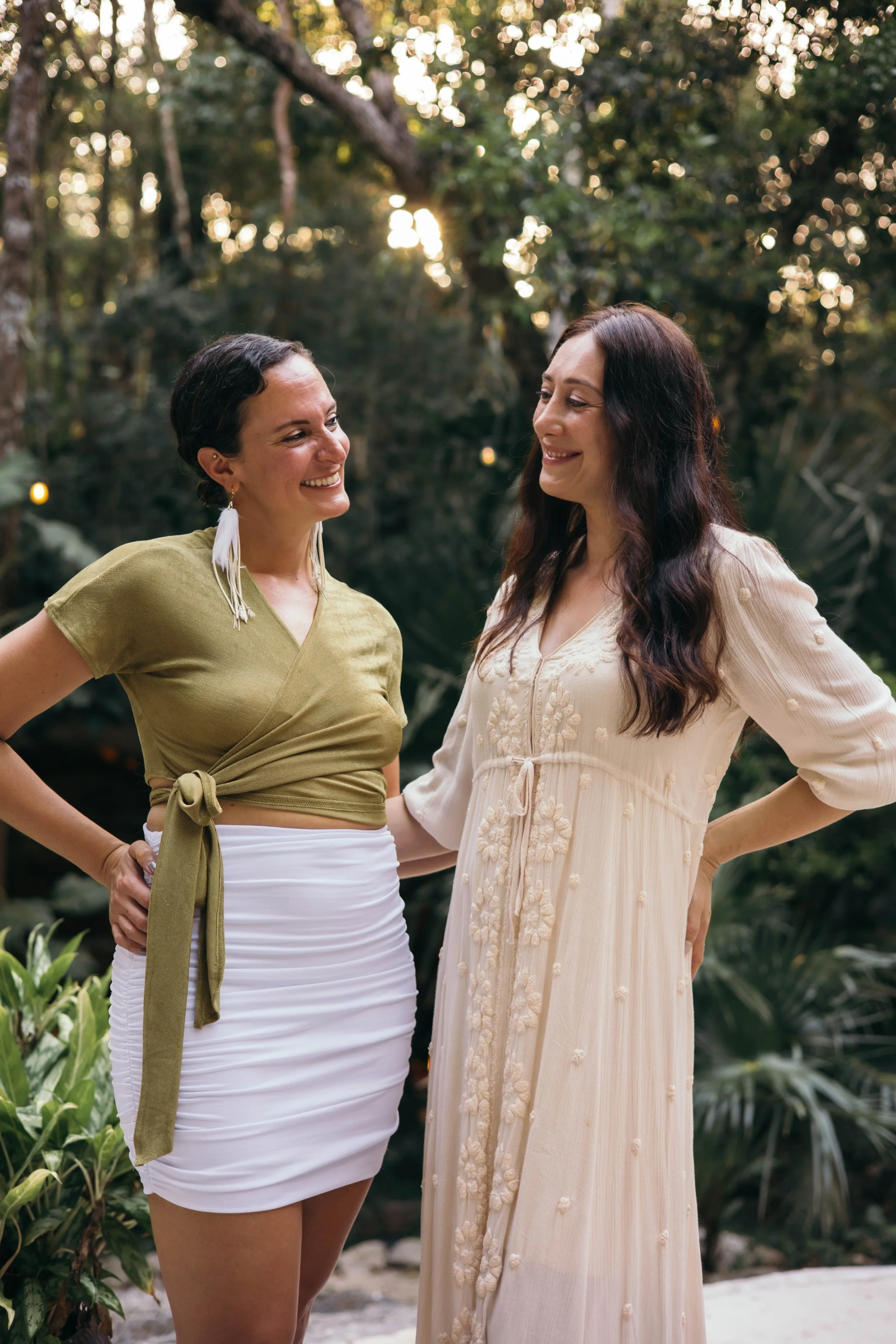 Two women smiling and talking in a lush, outdoor setting with trees and sunlight in the background.
