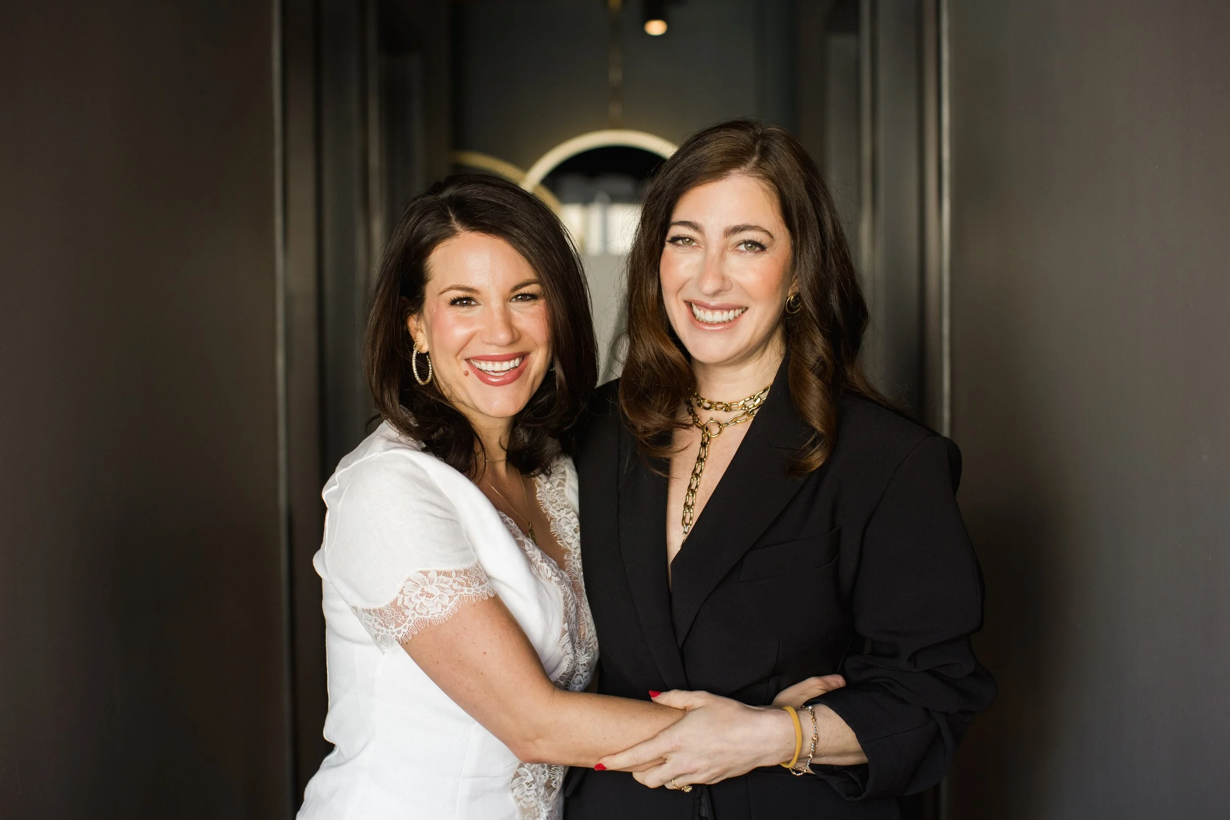 Two women standing close together, smiling and holding hands, in an indoor setting with dark walls and a round light fixture in the background.