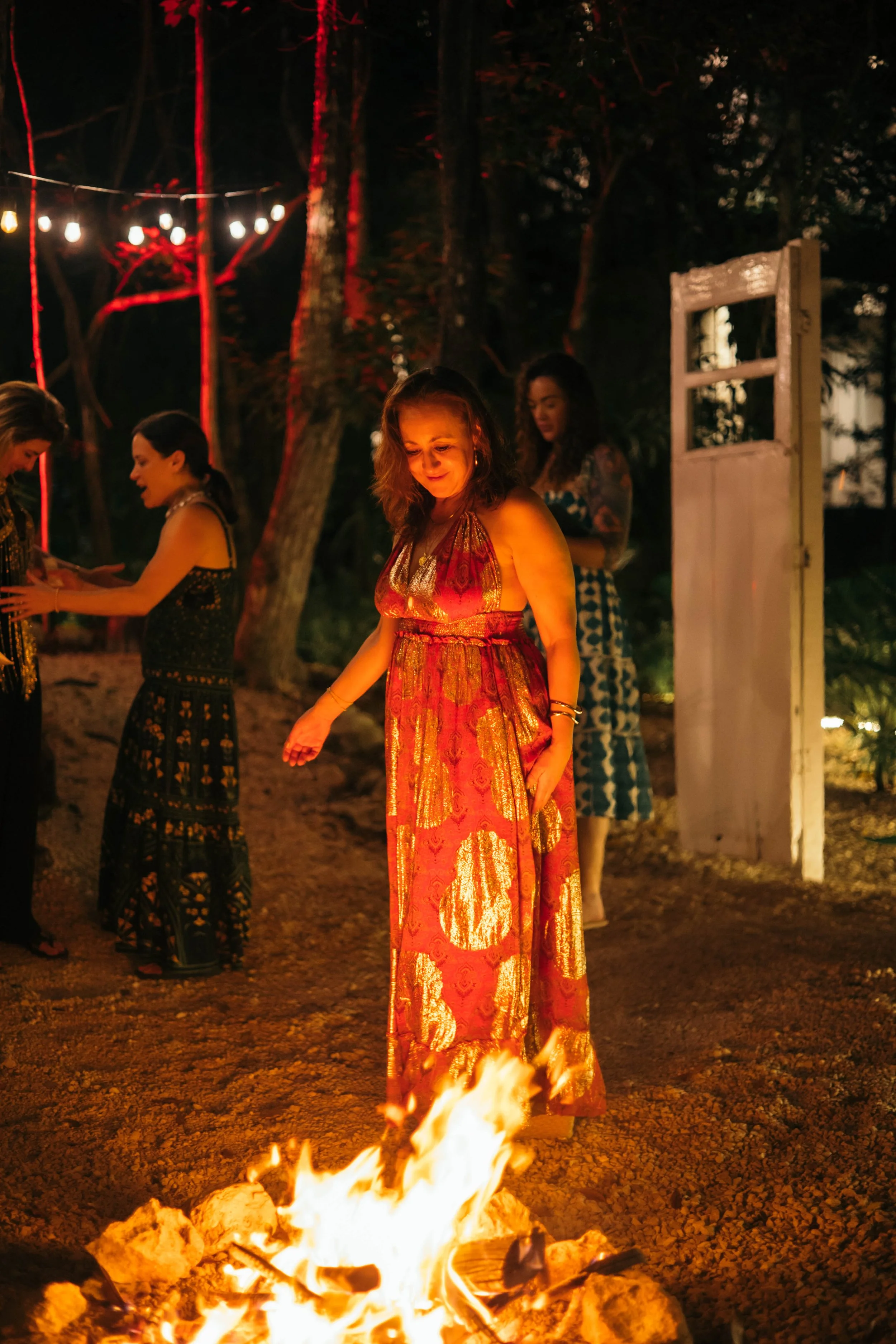People gathered around a bonfire at an outdoor evening event with string lights overhead.