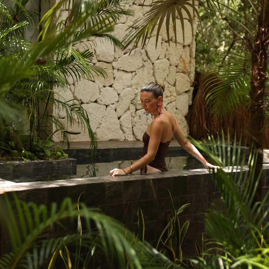 Woman leaning on the edge of a black stone bathtub surrounded by tropical plants and a stone wall.