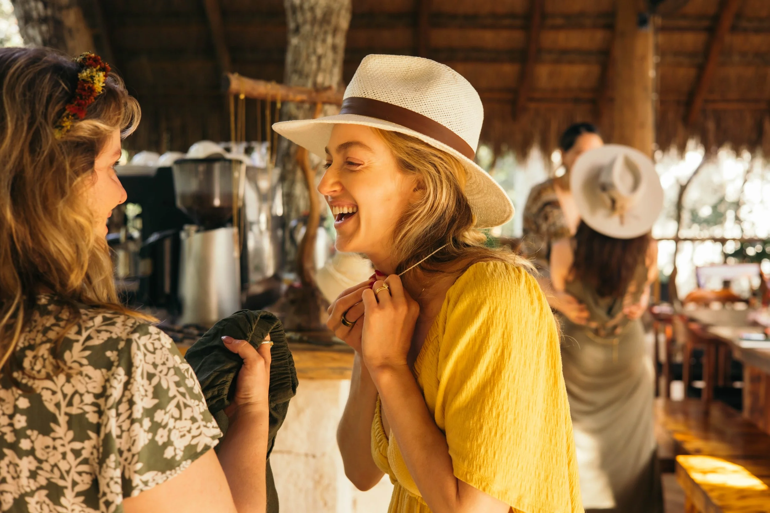 Two women smiling and conversing, one wearing a large sun hat and a yellow dress, the other with wavy hair and a flower headband, in a rustic outdoor setting with trees and wooden structures.