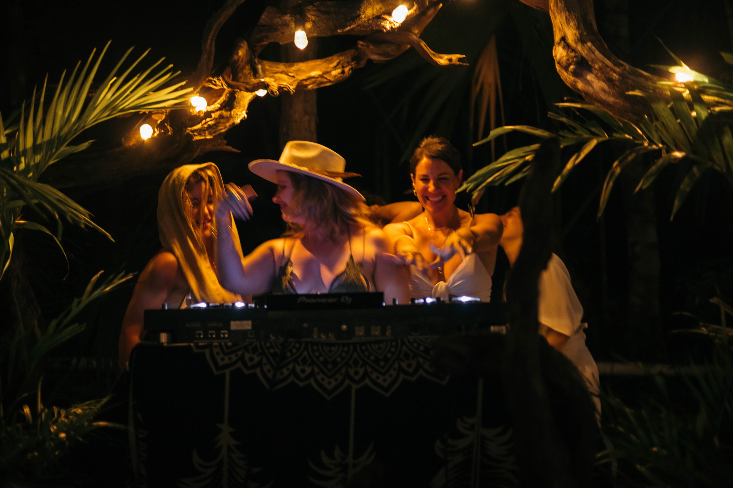 Three women dancing and smiling around a DJ booth at a tropical-themed party with dim lighting, lanterns, and palm leaves.