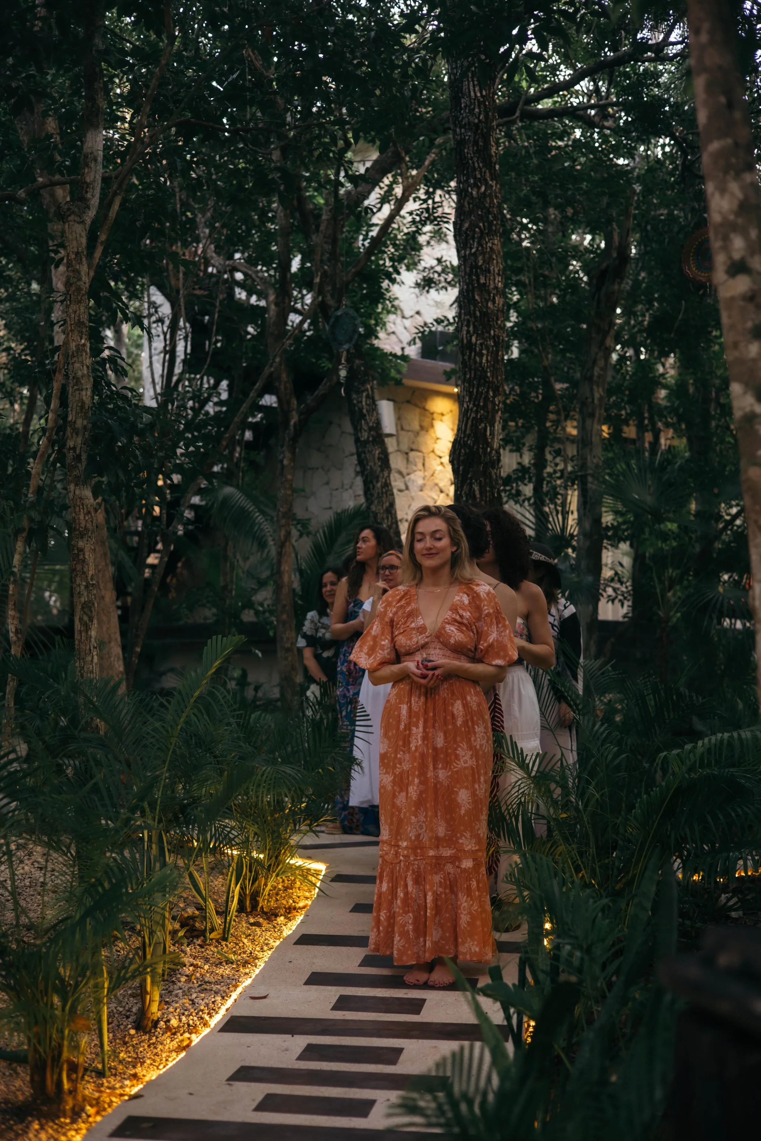 People standing in line outdoors on a pathway in a lush garden, with woman in an orange floral dress at the front.