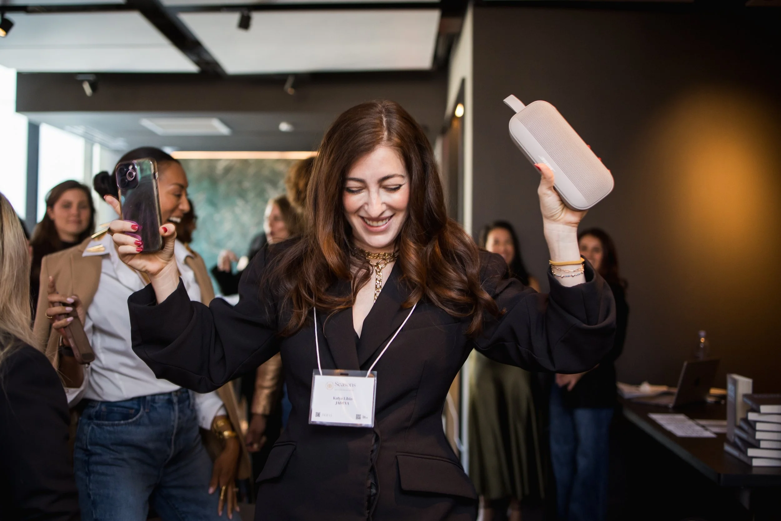 A woman dancing at a conference or event, holding a speaker in one hand and a smartphone in the other. She is smiling and wearing a black blazer with a conference badge.