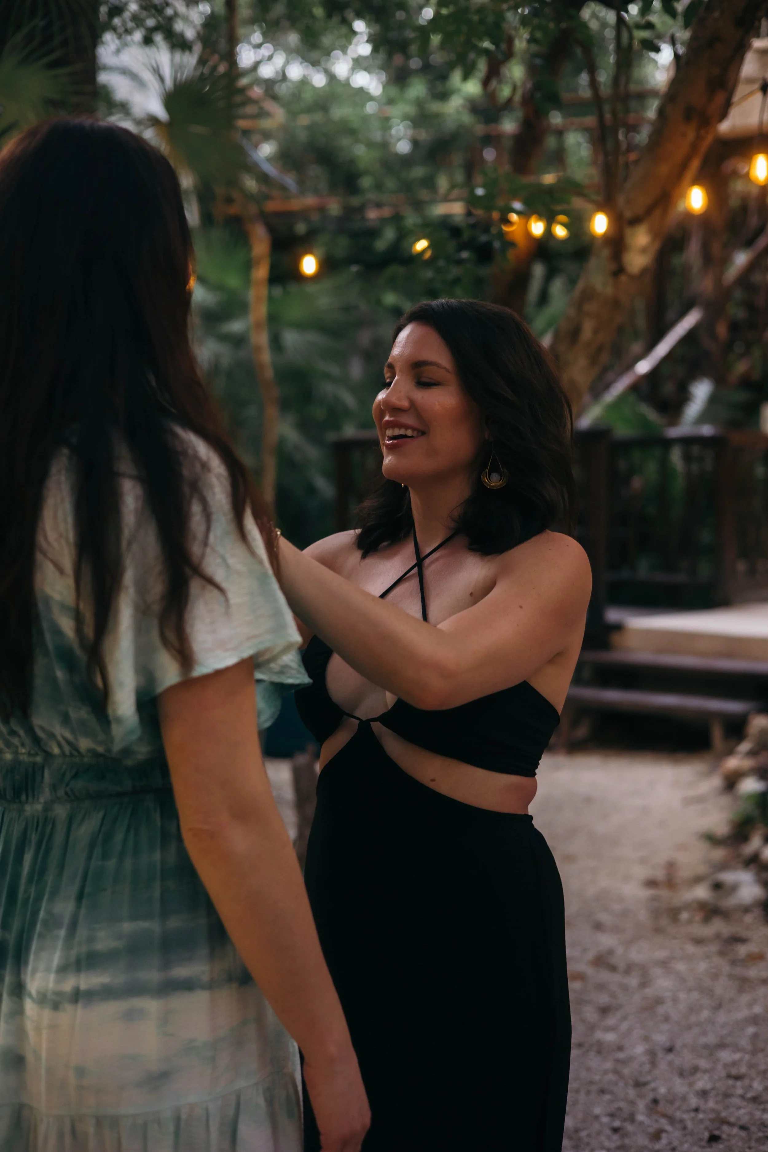 Two women standing outdoors in a wooded area at dusk, with string lights hanging in the background. One woman with dark, shoulder-length hair and earrings is smiling and touching the shoulder of the other woman, who has longer dark hair and is wearing a dress.