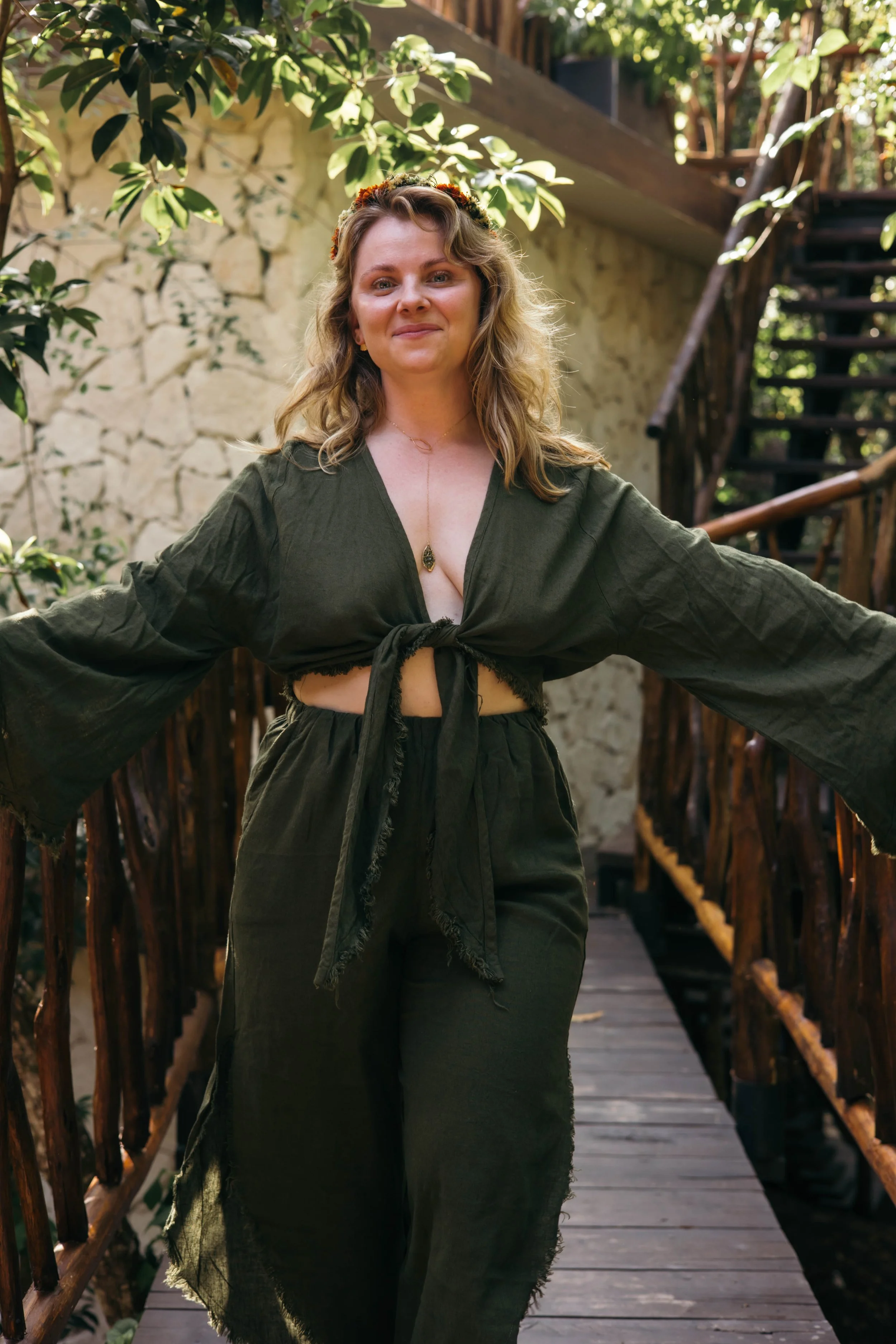 A woman standing on a wooden bridge outdoors, wearing a dark green outfit with a tied top and pants, with her arms outstretched and smiling at the camera.