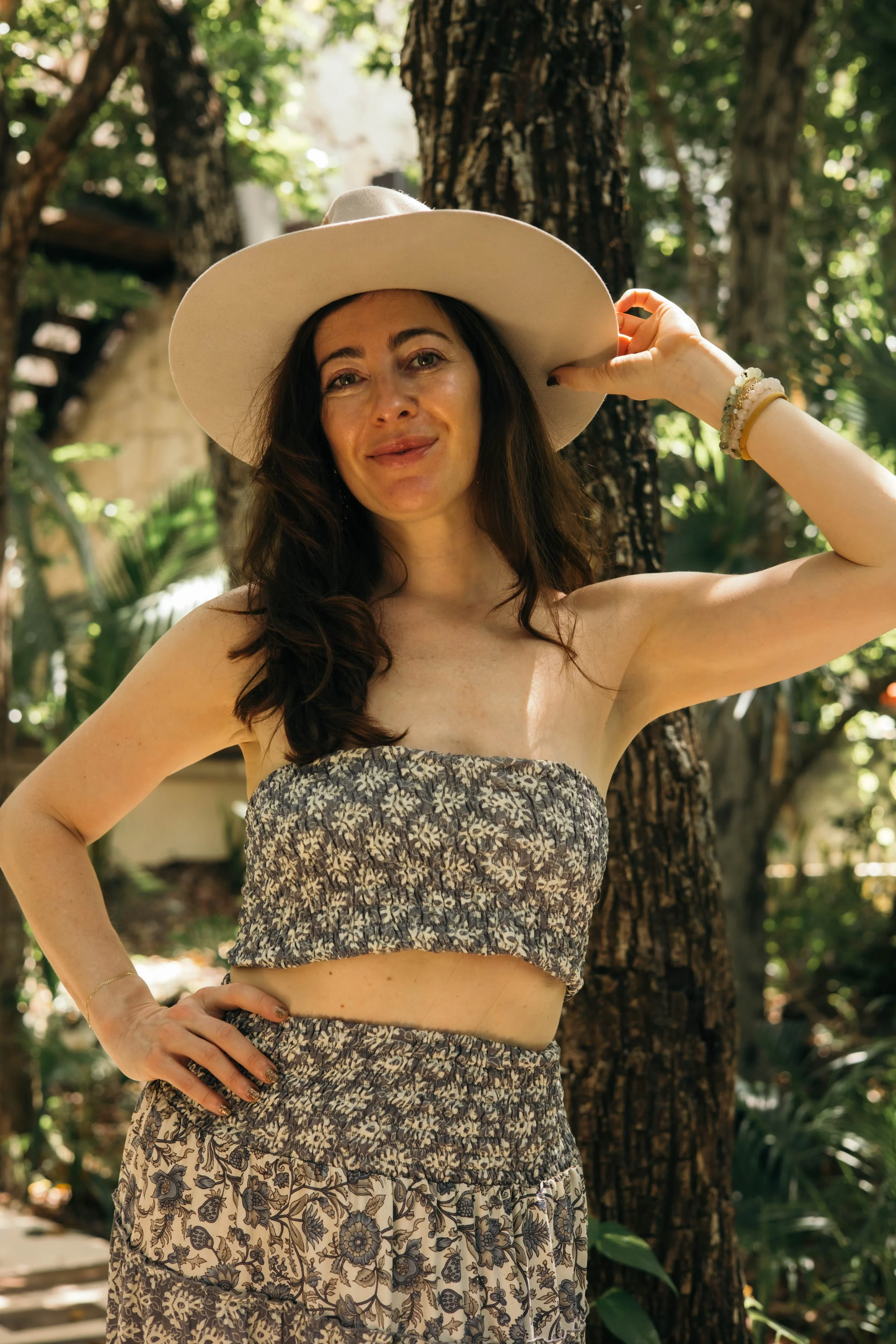 A woman with dark hair, wearing a wide-brimmed hat and a strapless floral outfit, standing outdoors in a wooded area.