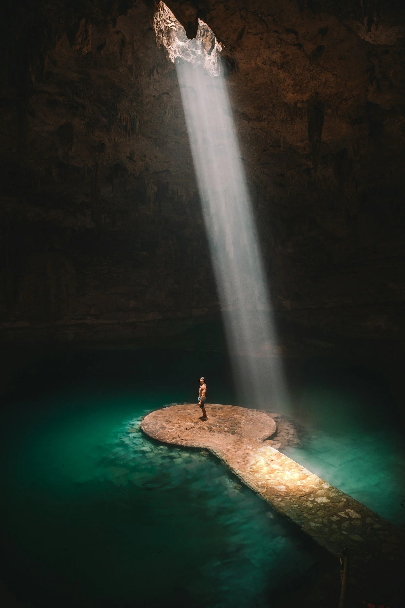 Person standing on a rock formation inside a dark cave with a waterfall and a beam of sunlight shining through an opening overhead, illuminating the water and the person.