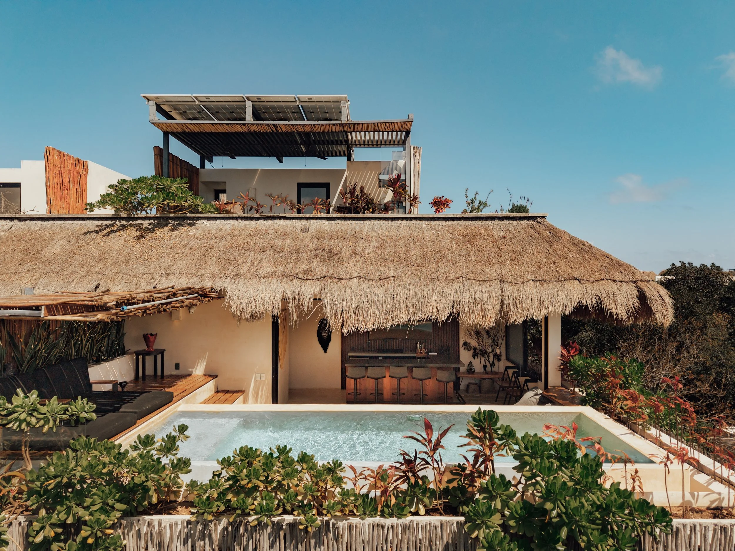 A modern house with a thatched roof, a rooftop deck with solar panels, an outdoor pool, and surrounding lush greenery under a clear blue sky.