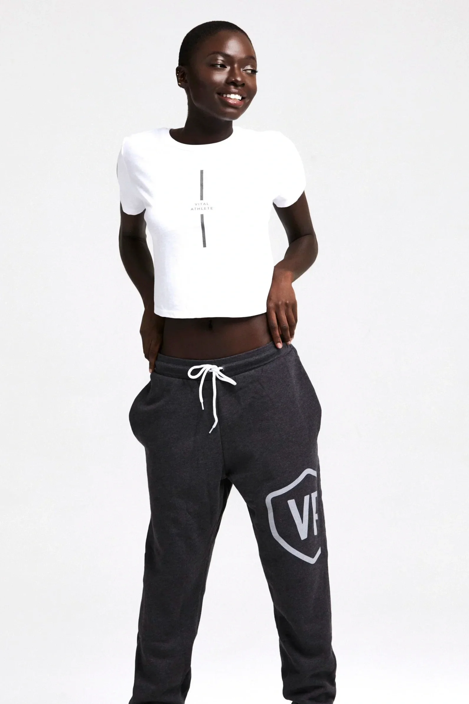 A woman standing against a plain white wall, wearing a white t-shirt with text and graphics and dark sweatpants with a logo, smiling and relaxed.