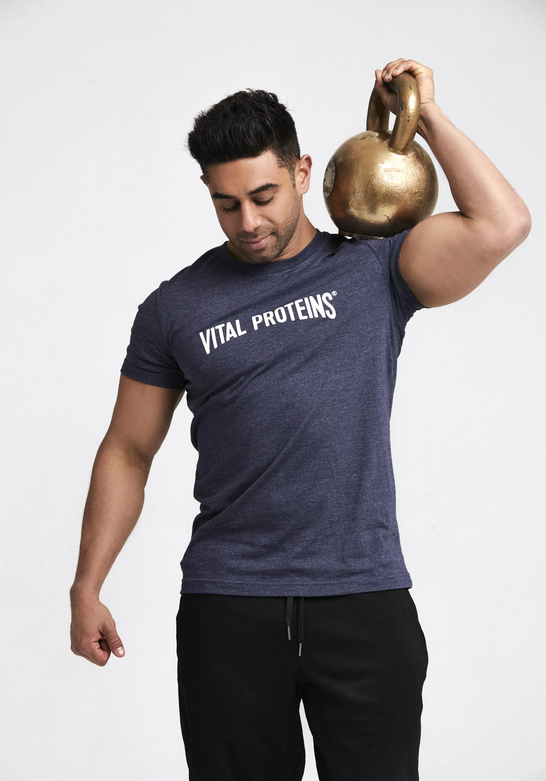 A man lifting a golden kettlebell on his shoulder, wearing a navy blue shirt with 'VITAL PROTEINS' written on it, against a plain white background.