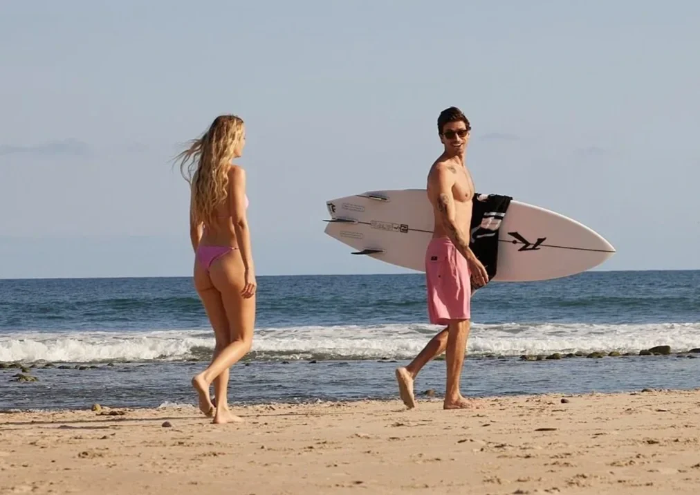 A man and woman walking on the beach near the ocean. The man is holding a surfboard and is wearing pink shorts. The woman has long blonde hair and is wearing a pink bikini.