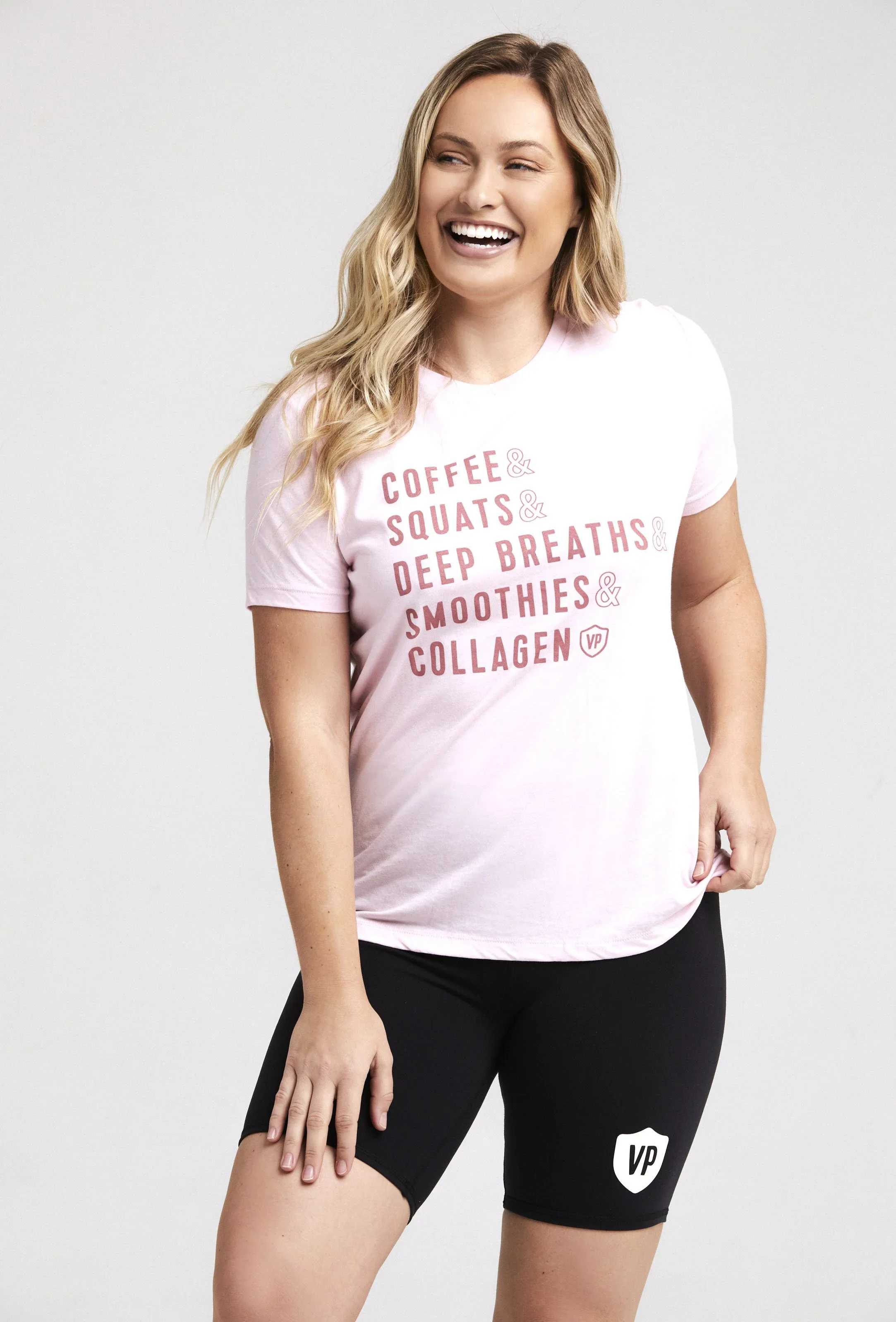 A woman wearing a pink t-shirt with fitness-related words and black shorts, smiling and touching her leg against a plain gray background.