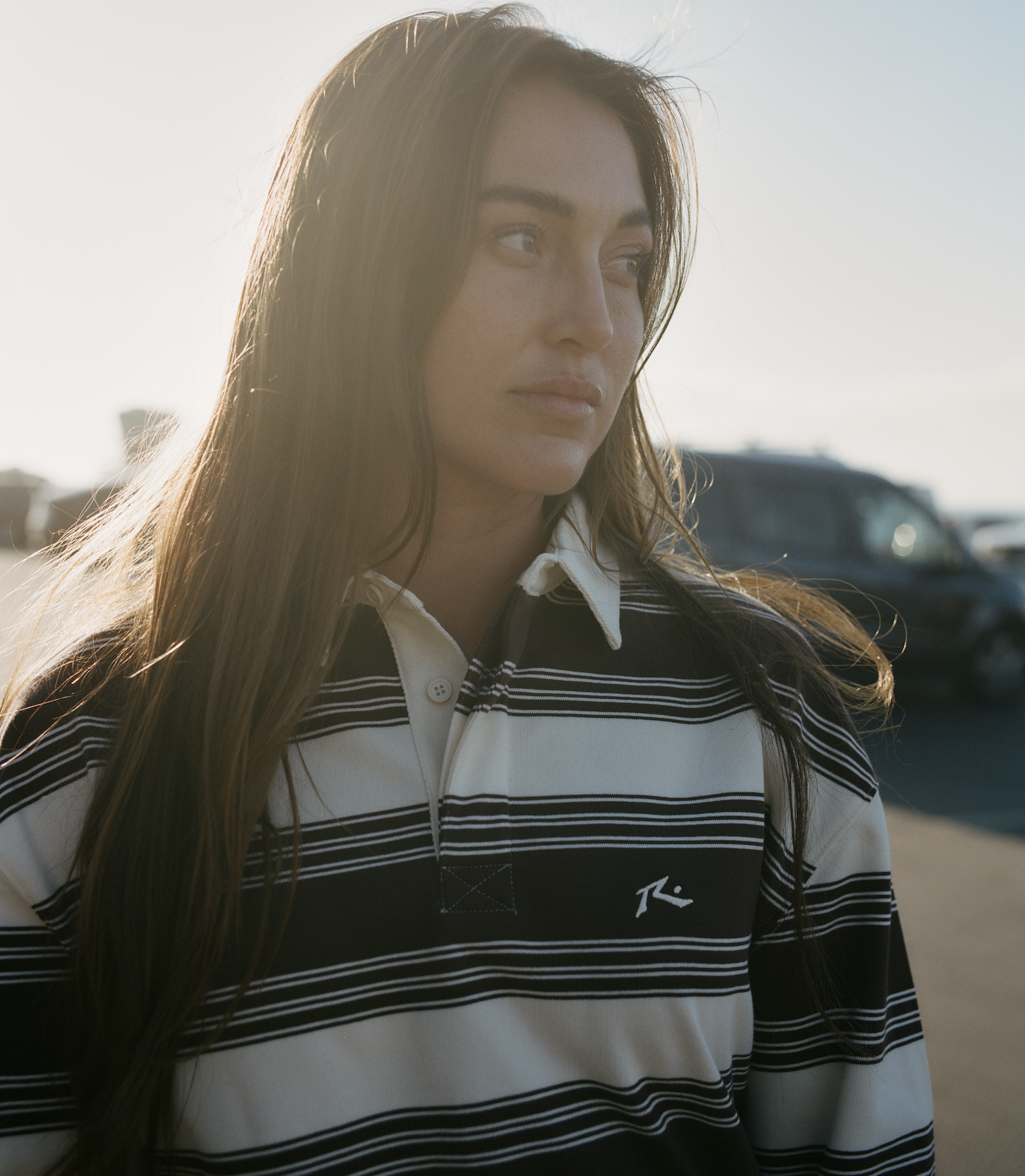 A young woman with long brown hair standing outdoors in front of a parking lot full of cars, backlit by sunlight, wearing a striped rugby shirt.