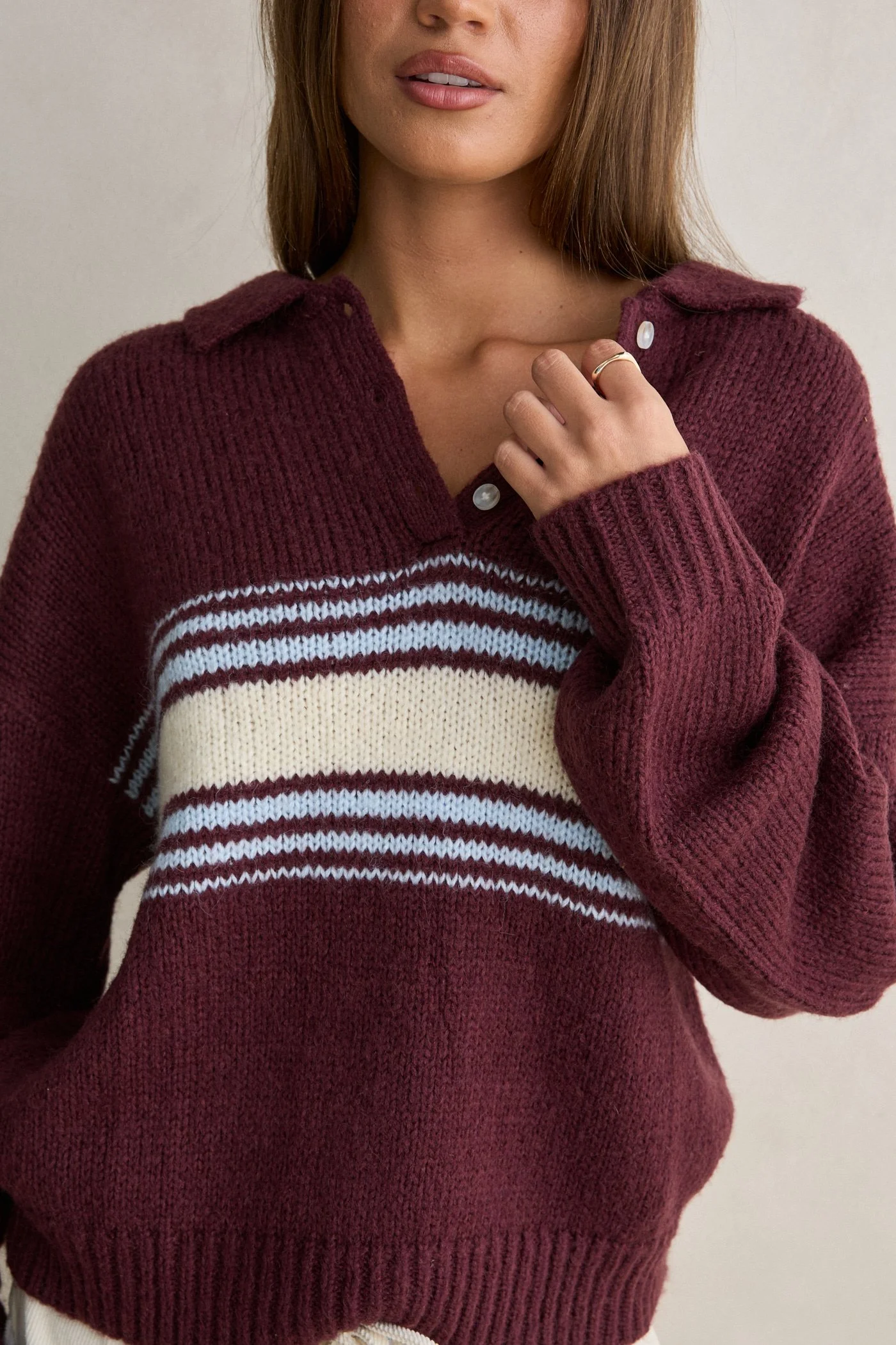 Close-up of a woman wearing a maroon knit sweater with white stripes, touching her neck with one hand, with a neutral background.