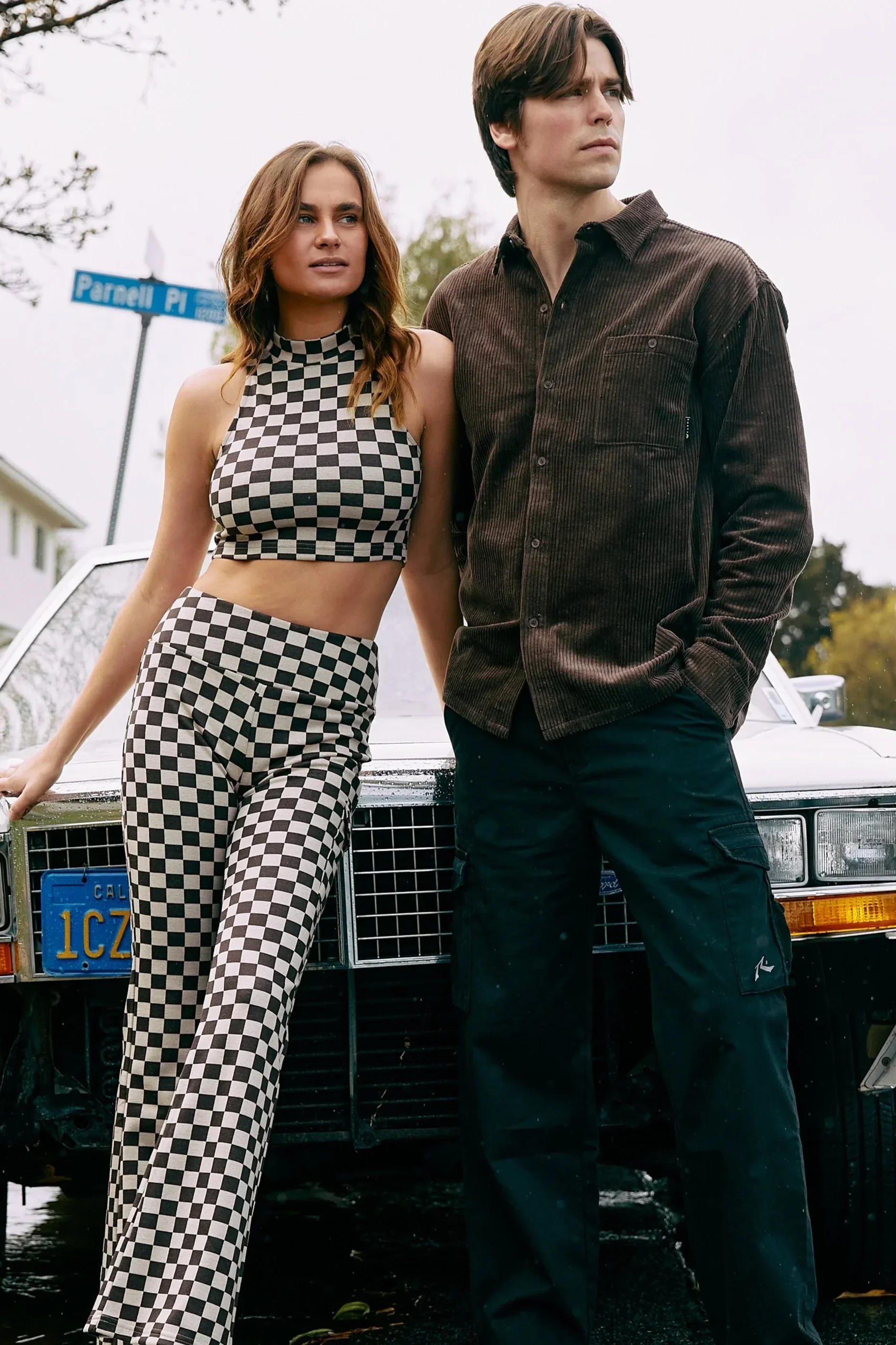 A woman in checkered pants and top stands next to a man in dark clothes, both leaning against a vintage car with a California license plate. They are outdoors, with a street sign in the background that reads Parcells Pi.