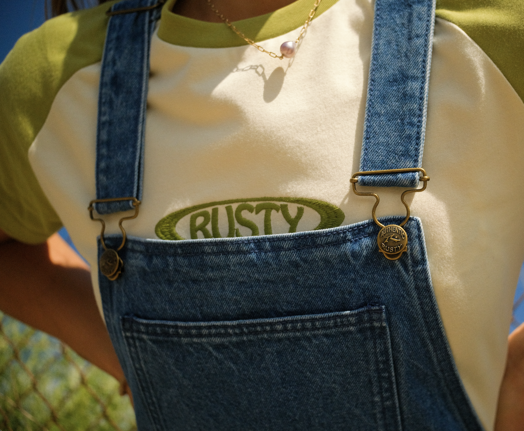 Close-up of a person's torso wearing a yellow shirt with a green logo that says 'Rusty,' denim overalls, a gold chain necklace with a pink pearl, and a gold button labeled 'Authentic Rusty.'