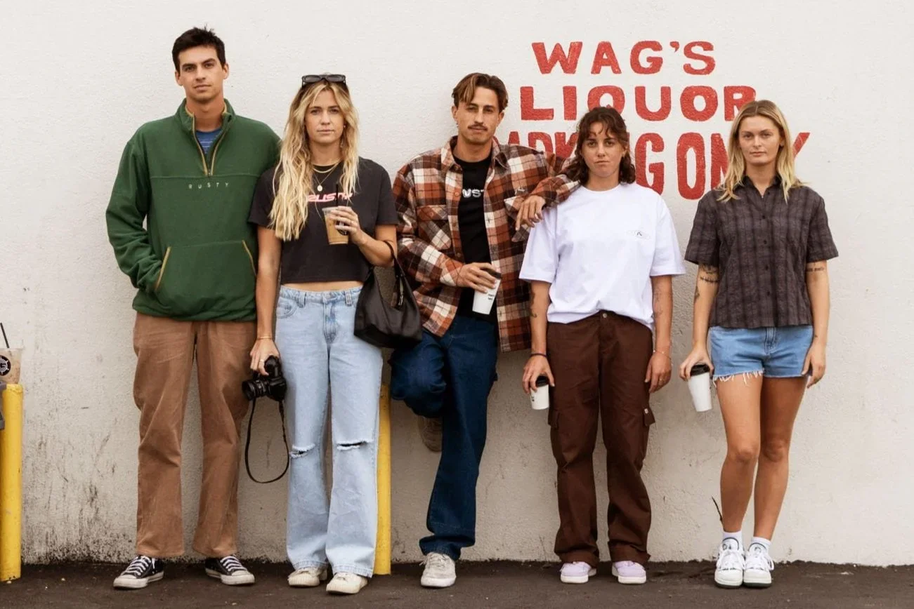 Five young adults standing against a white wall with red text that reads "WAG'S LIQUOR ADVENTURE". They are casually dressed, some holding drinks and a camera.