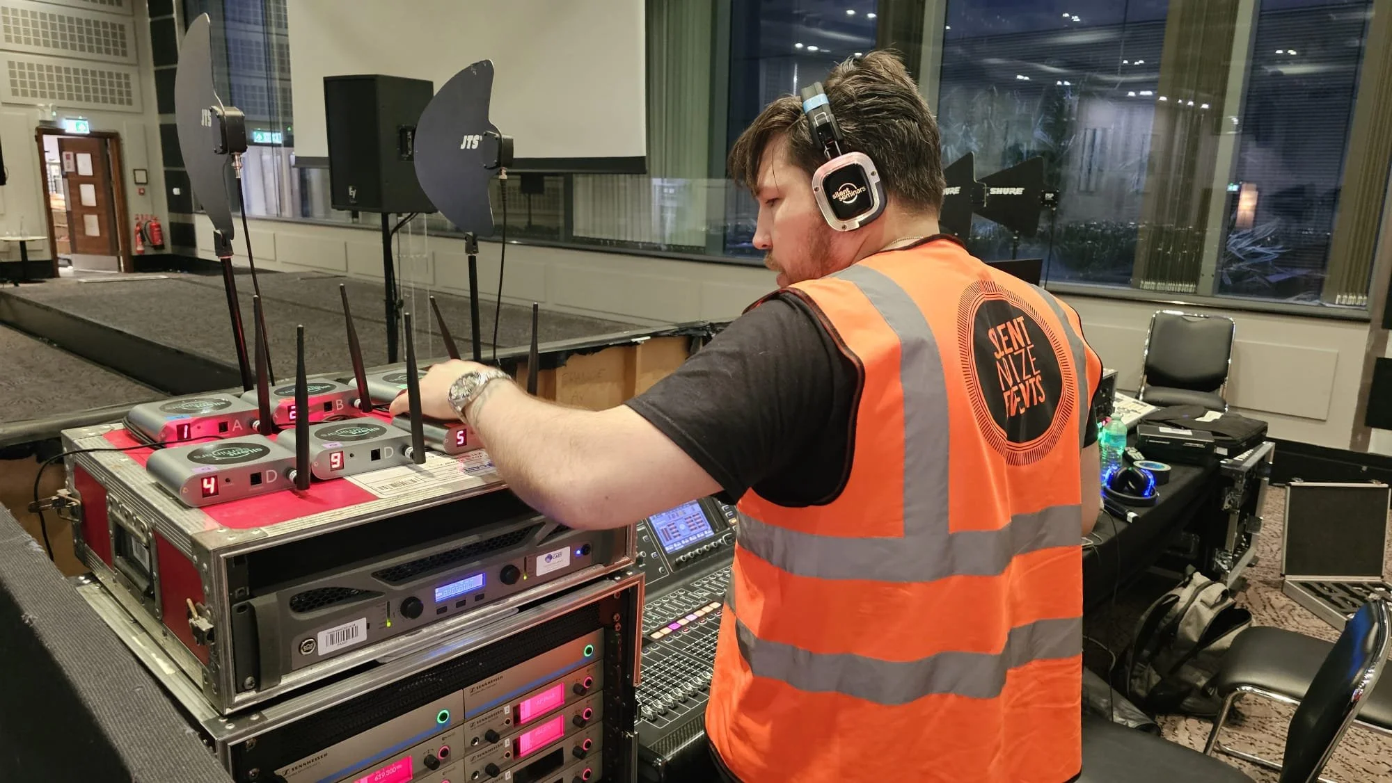 A man wearing a black shirt, orange safety vest, and headphones is adjusting equipment on a sound and lighting control table at an indoor event. The vest has the logo 'Sent Mize Events' on the back. In the background, there are large windows and sound barriers.