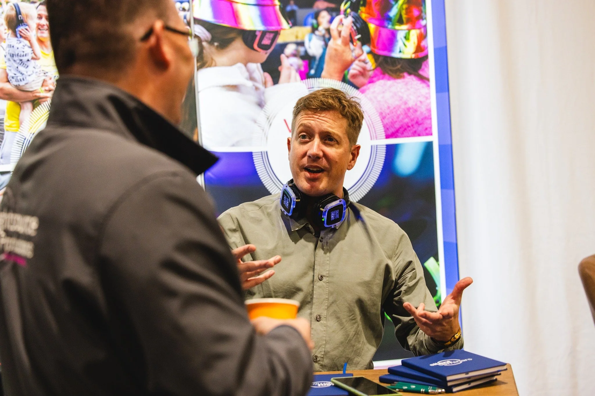 Two men engaged in conversation at a booth, with one man facing the camera wearing headphones around his neck. There are notebooks, pens, and a phone on the table, with a colorful promotional display in the background.
