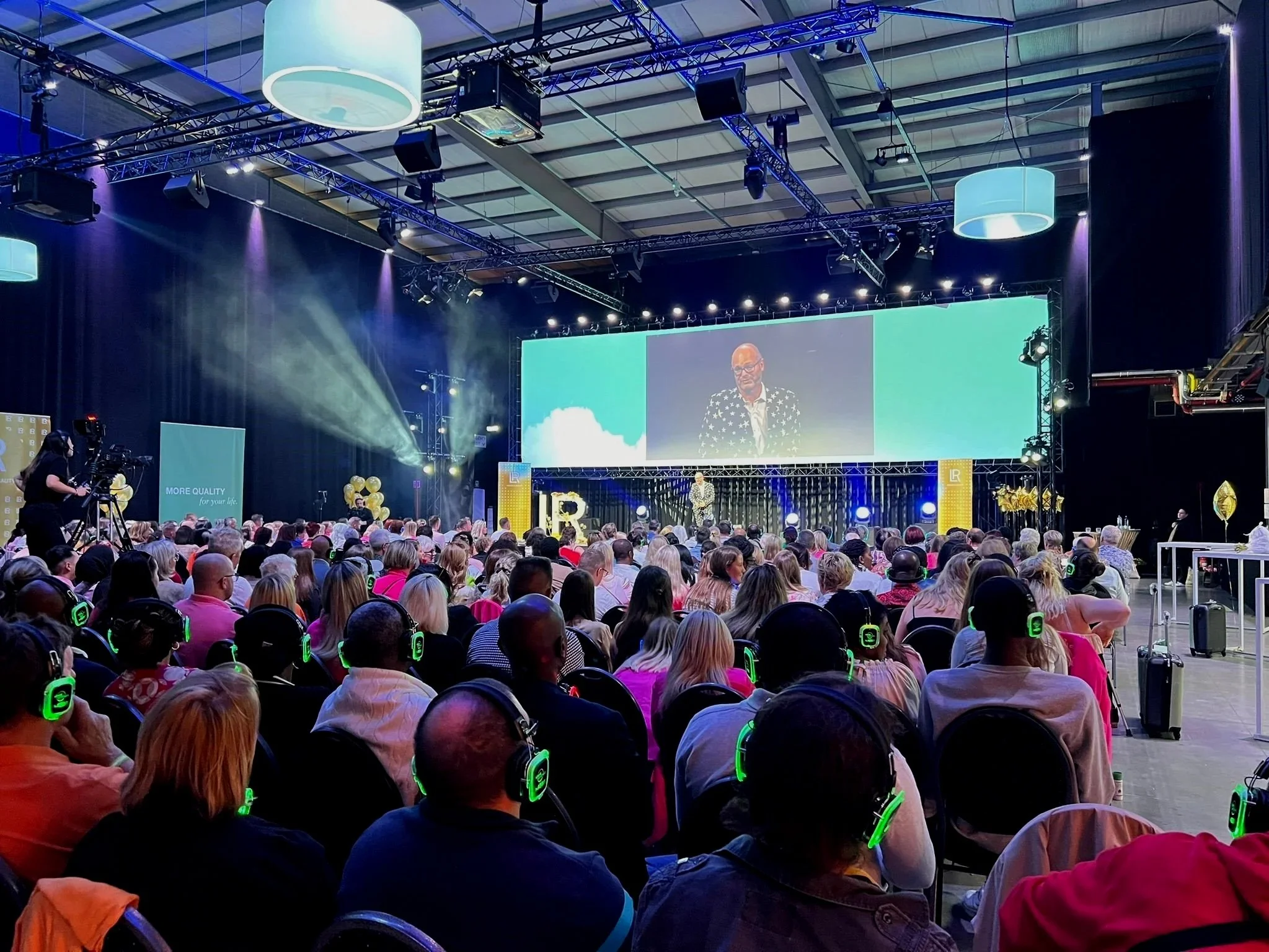 Large audience wearing green headphones attending a conference or presentation in a spacious hall with a stage and large screen displaying a speaker in a polka dot blazer.