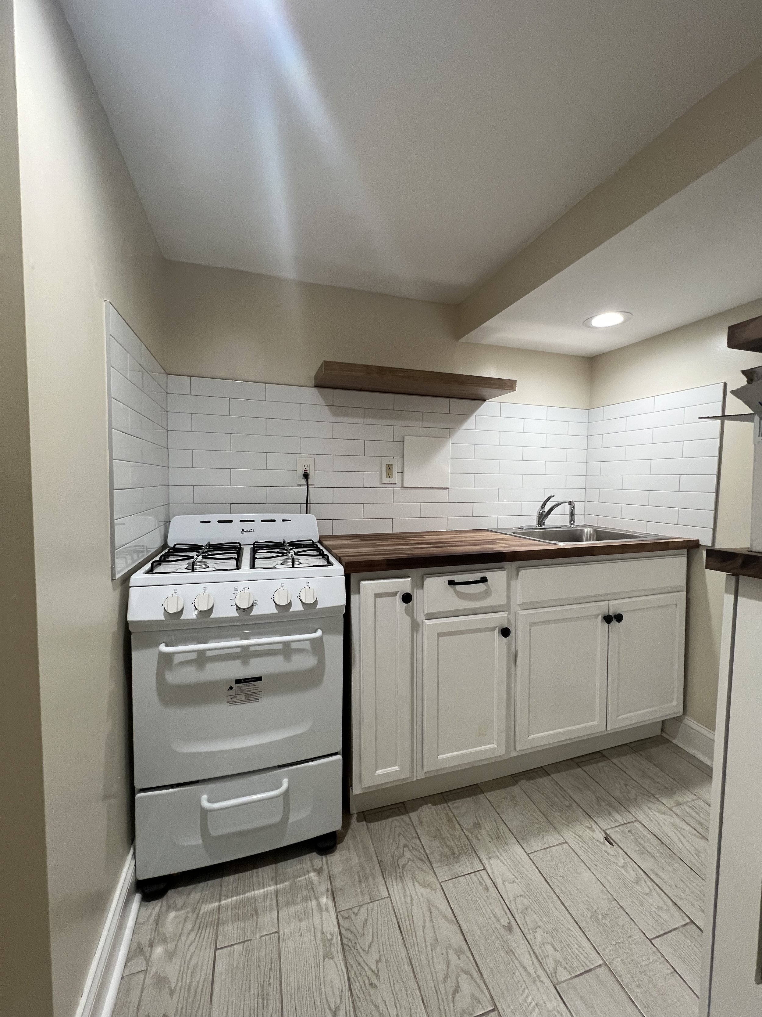 Small kitchen with white cabinets, wooden countertop, white stove, and white tiled backsplash, including a sink and shelving.