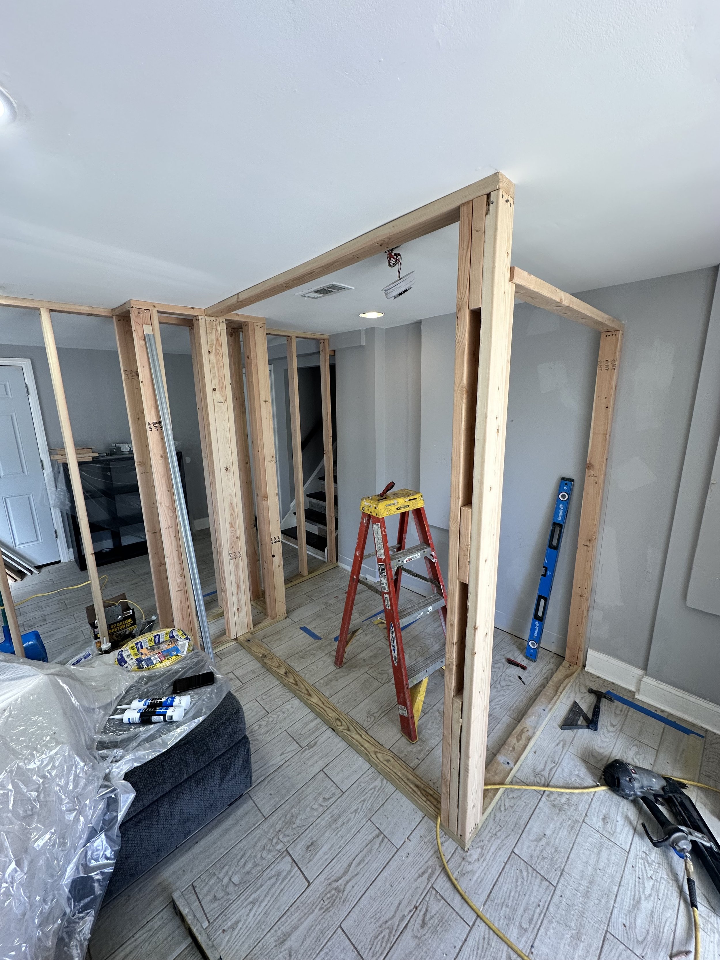 Interior view of a room under construction with wooden framing partially built, construction tools, a red ladder, and a blue level.