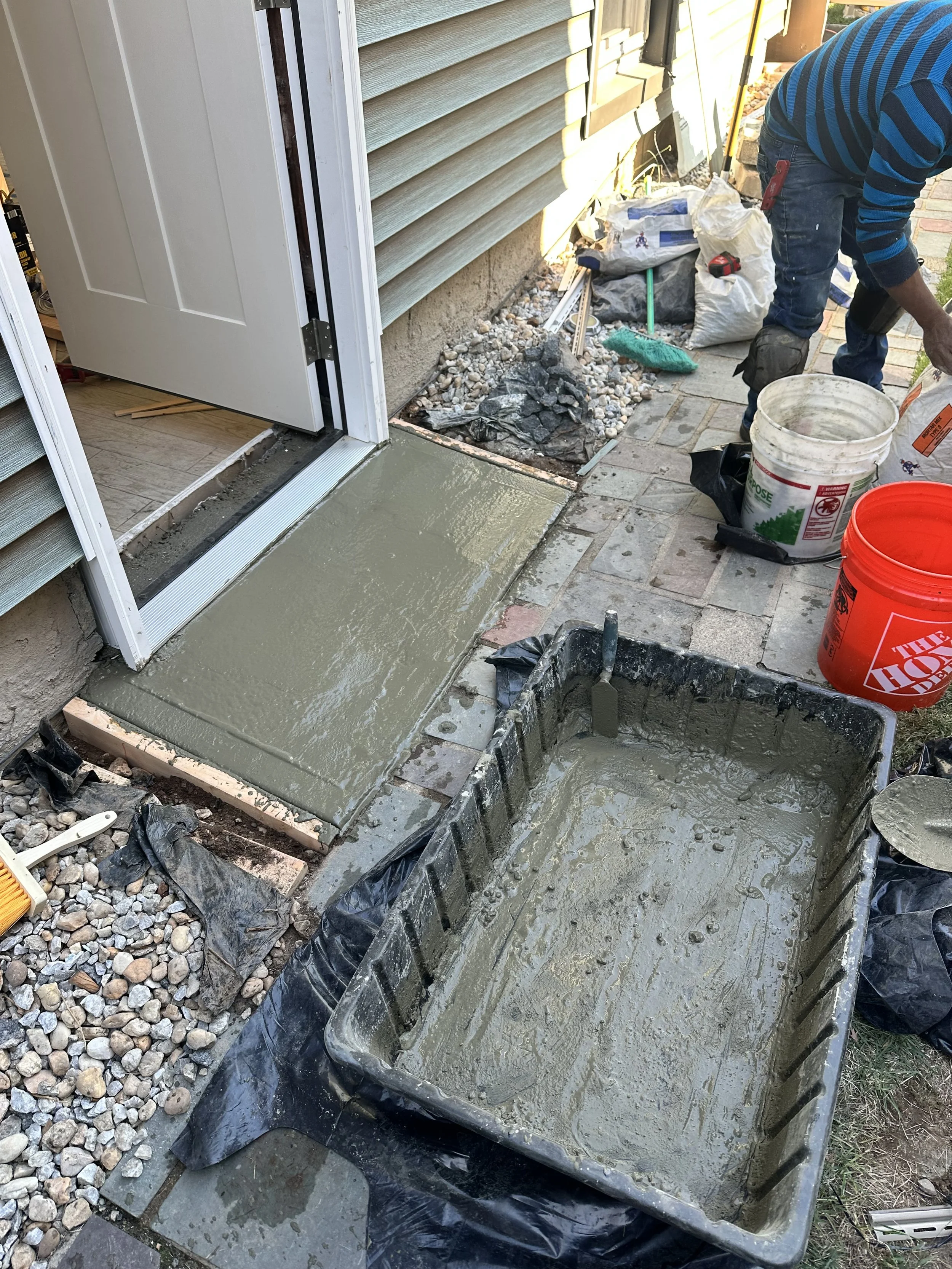A person working on a concrete step at a doorway, with tools and buckets filled with concrete around. The step is freshly poured and still wet.