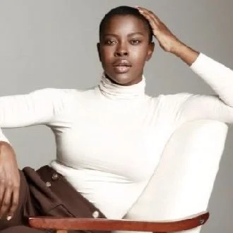 A person sitting on a chair wearing a white turtleneck and brown pants, posing with one hand on their head and the other resting on the chair arm. The background is neutral.