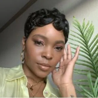 Person with short hair wearing a green shirt and gold hoop earrings in front of a plant.
