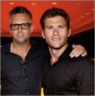 Two men posing for a photo together in a dimly lit setting, both wearing black shirts, with one man having glasses and short hair and the other having a styled haircut. The background has a warm, orange-red tone.