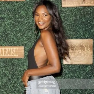 Person posing in front of a greenery wall at an event, wearing a black top and jeans.