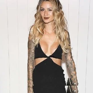 Person in a black cutout dress with tattooed arms standing against a white background.