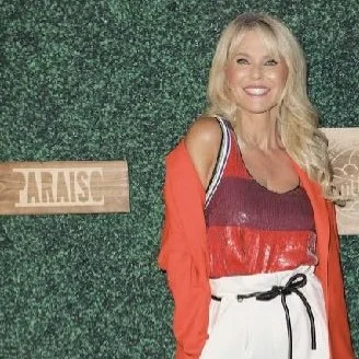 Woman posing in front of a green backdrop with wooden sign "PARAISO," wearing a red top, white pants, and a red jacket.