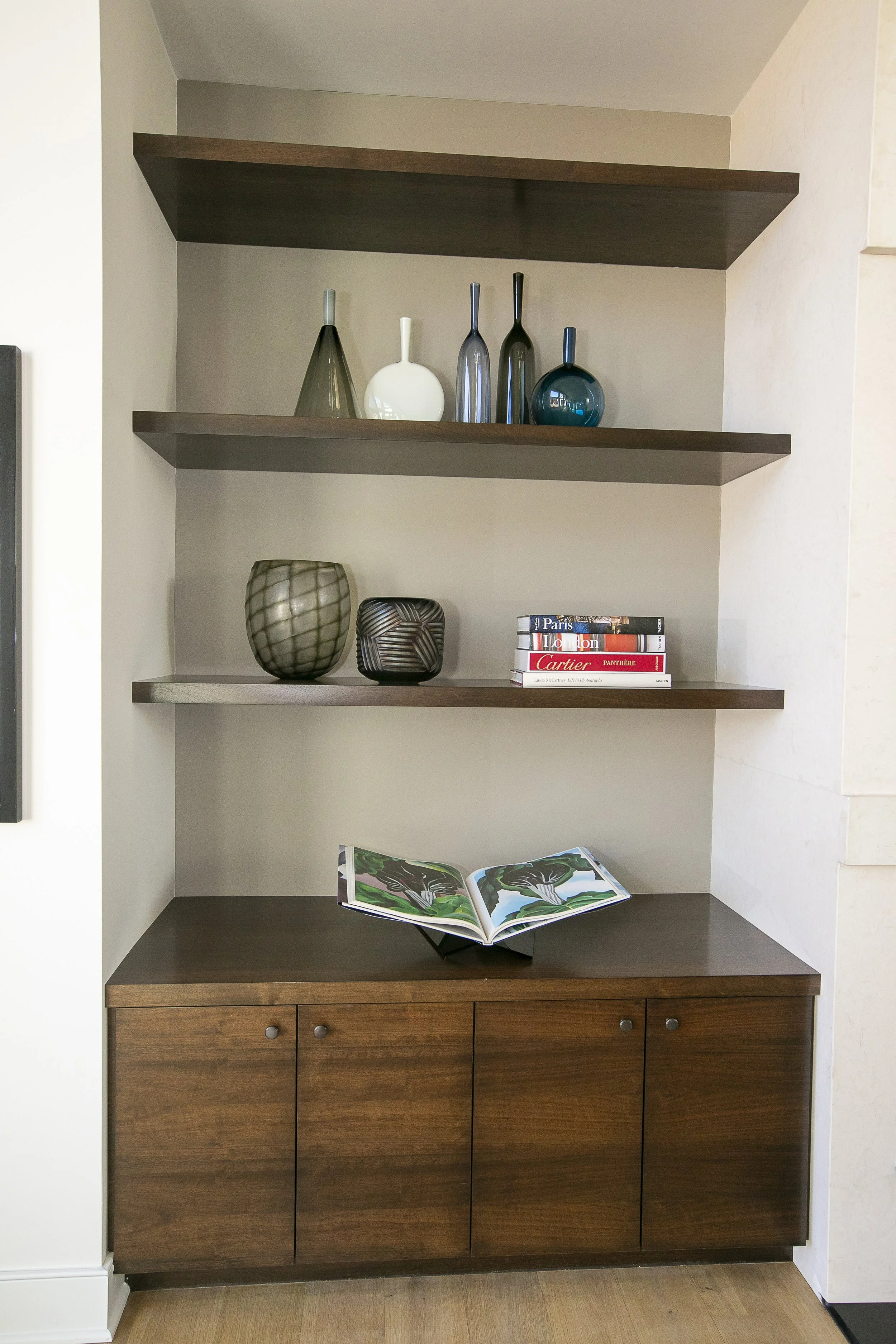 Various vases in clear, green, and blue glass add subtle color and texture on the shelves, while other sculptural decor pieces enhance the room’s modern aesthetic.