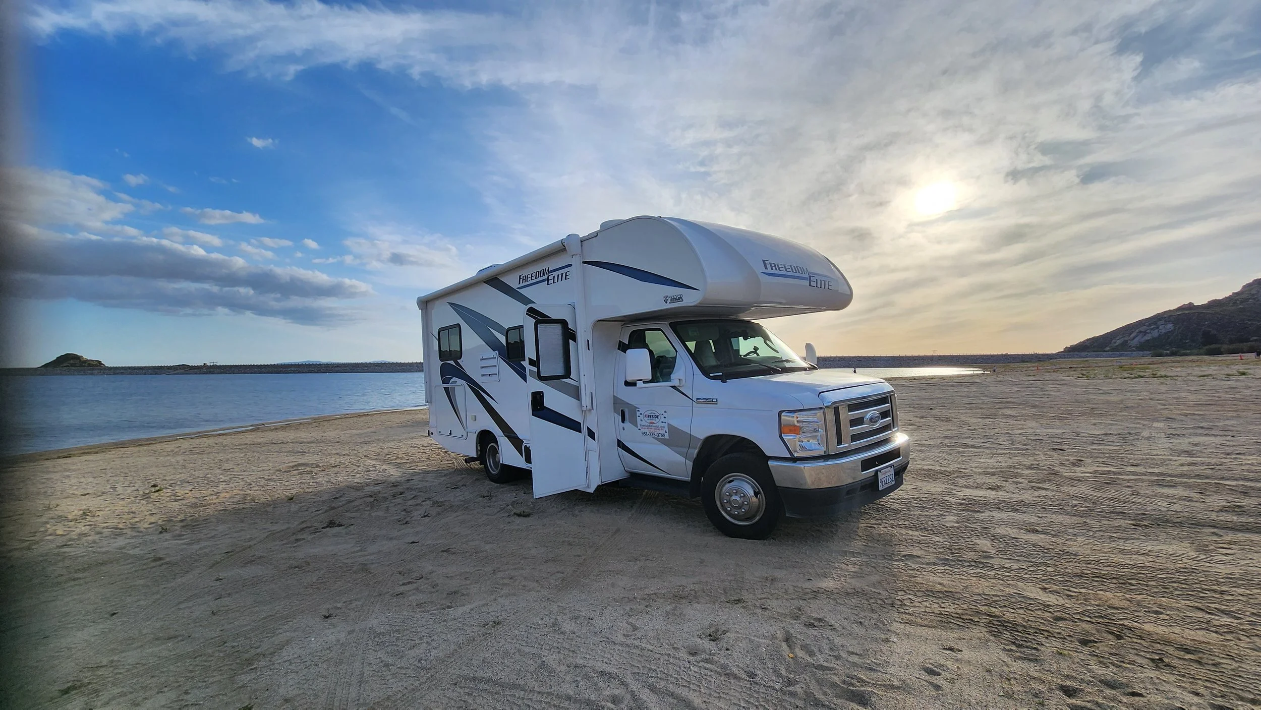 A white motorhome parked on a sandy beach by a body of water with hills in the background, under a partly cloudy sky during sunset.
