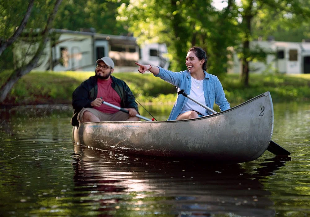 A woman and a man are canoeing on a calm lake surrounded by green trees, with RVs in the background; the woman is smiling and pointing while the man looks on.