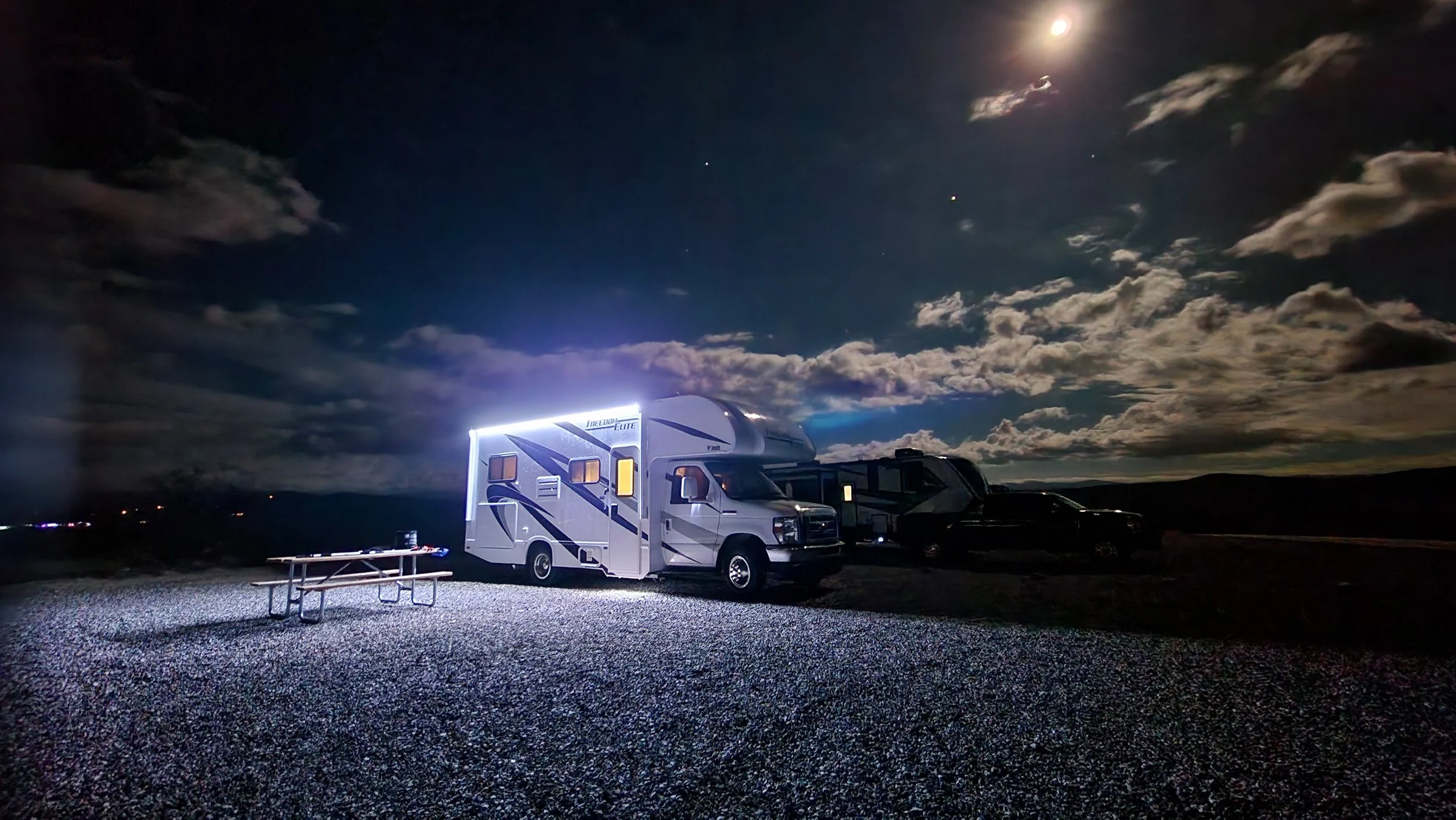 A recreational vehicle (RV) parked outdoors at night with a picnic table nearby. The sky is partly cloudy with the moon and stars visible, casting a glow over the scene.