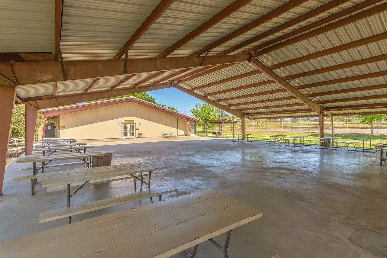 Empty outdoor pavilion with picnic tables, a building in the background, and a baseball field visible beyond the pavilion.
