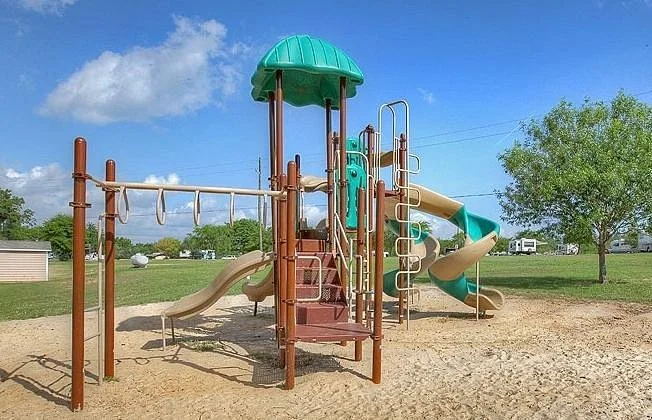 Children's playground with a brown and teal slide structure, climbing ladder, and swings, set on sandy ground in a park with green grass, trees, and a blue sky with clouds.
