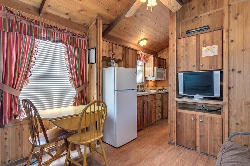Wood-paneled kitchen and dining area with striped curtains, a small dining table with three chairs, a white refrigerator, a microwave, a stove, a small television, and wooden cabinets.