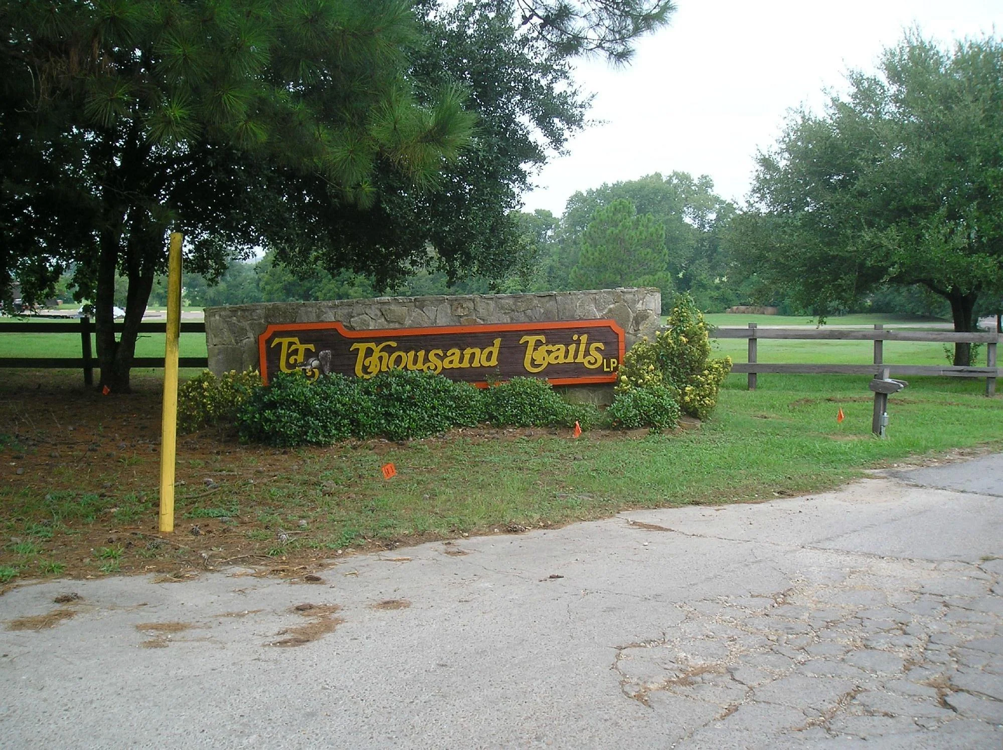 Entrance sign for Tear Thousand Trails with surrounding bushes, trees, grassy area, and partially visible road.