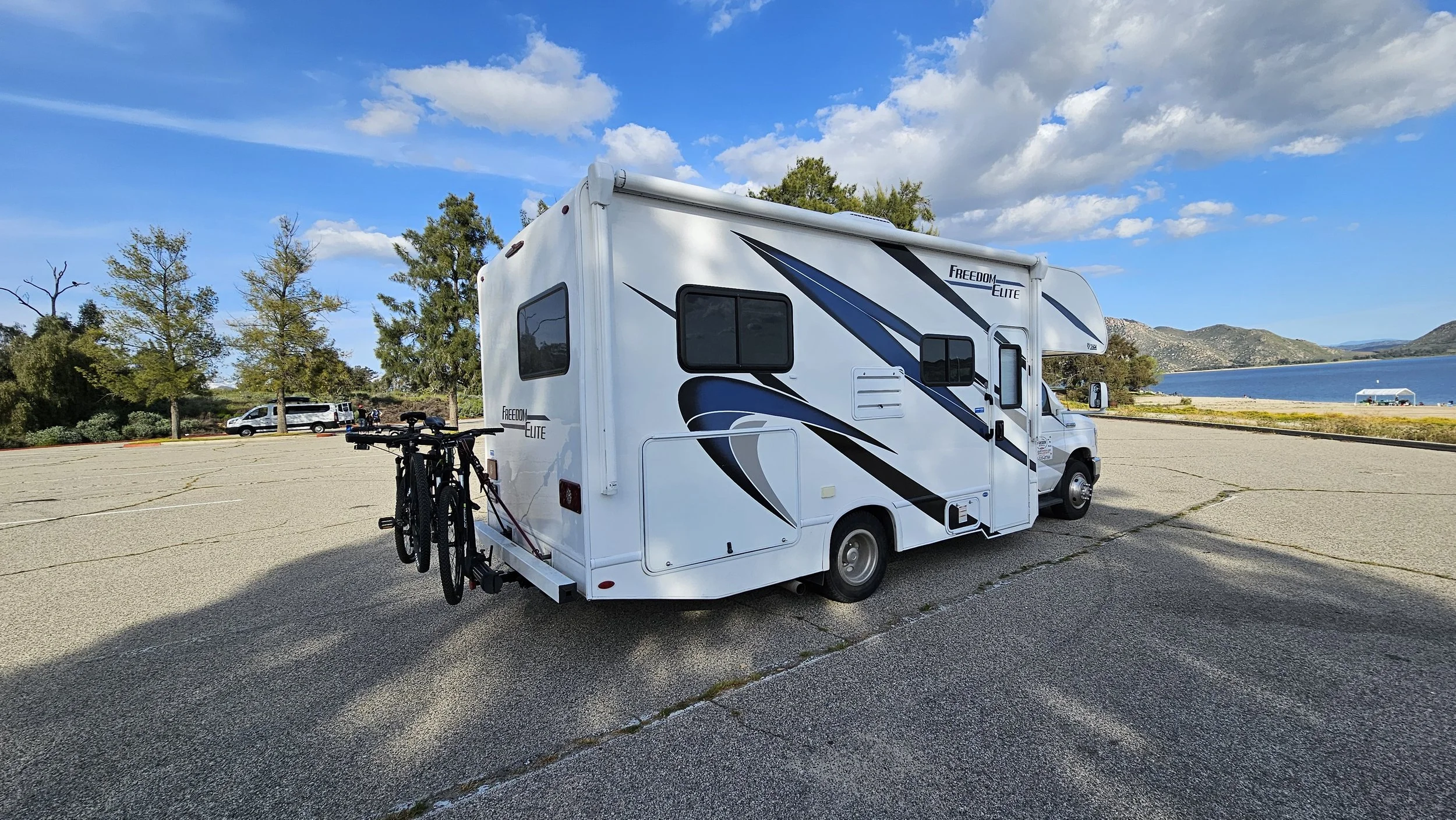 White RV with blue and black graphics parked in a lot near a lake, with bikes attached at the back, trees, hills, and a building in the background.