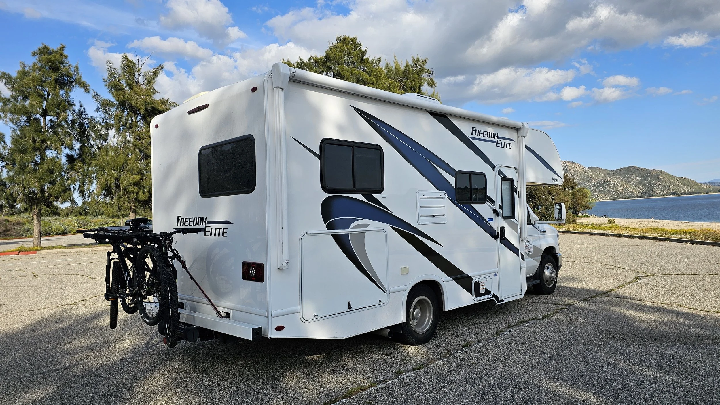White RV with black and blue graphics parked near a lake, with bike rack carrying two bikes on the back, trees on the left, mountains and water in the background, under a partly cloudy sky.