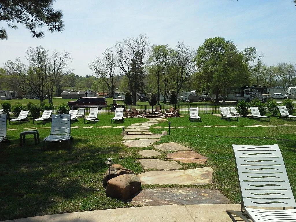Outdoor area with a winding stone pathway, lounge chairs arranged on the grass, trees with some in full leaf, a row of parked recreational vehicles, and a cloudy sky.