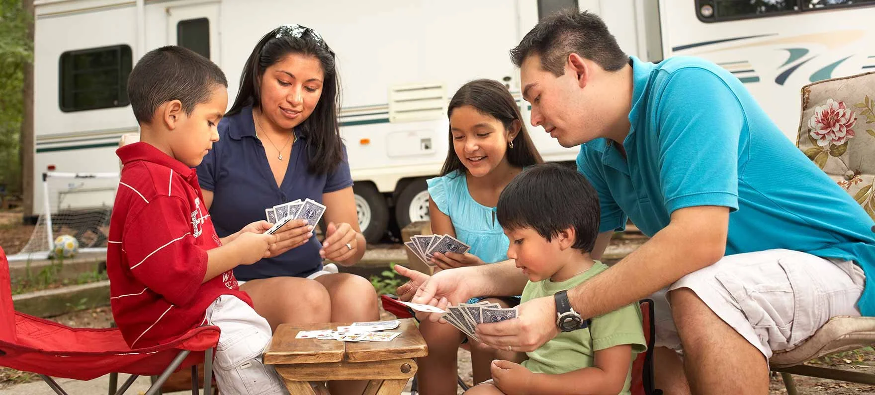 A family of five playing cards outdoors in a campsite with an RV in the background.