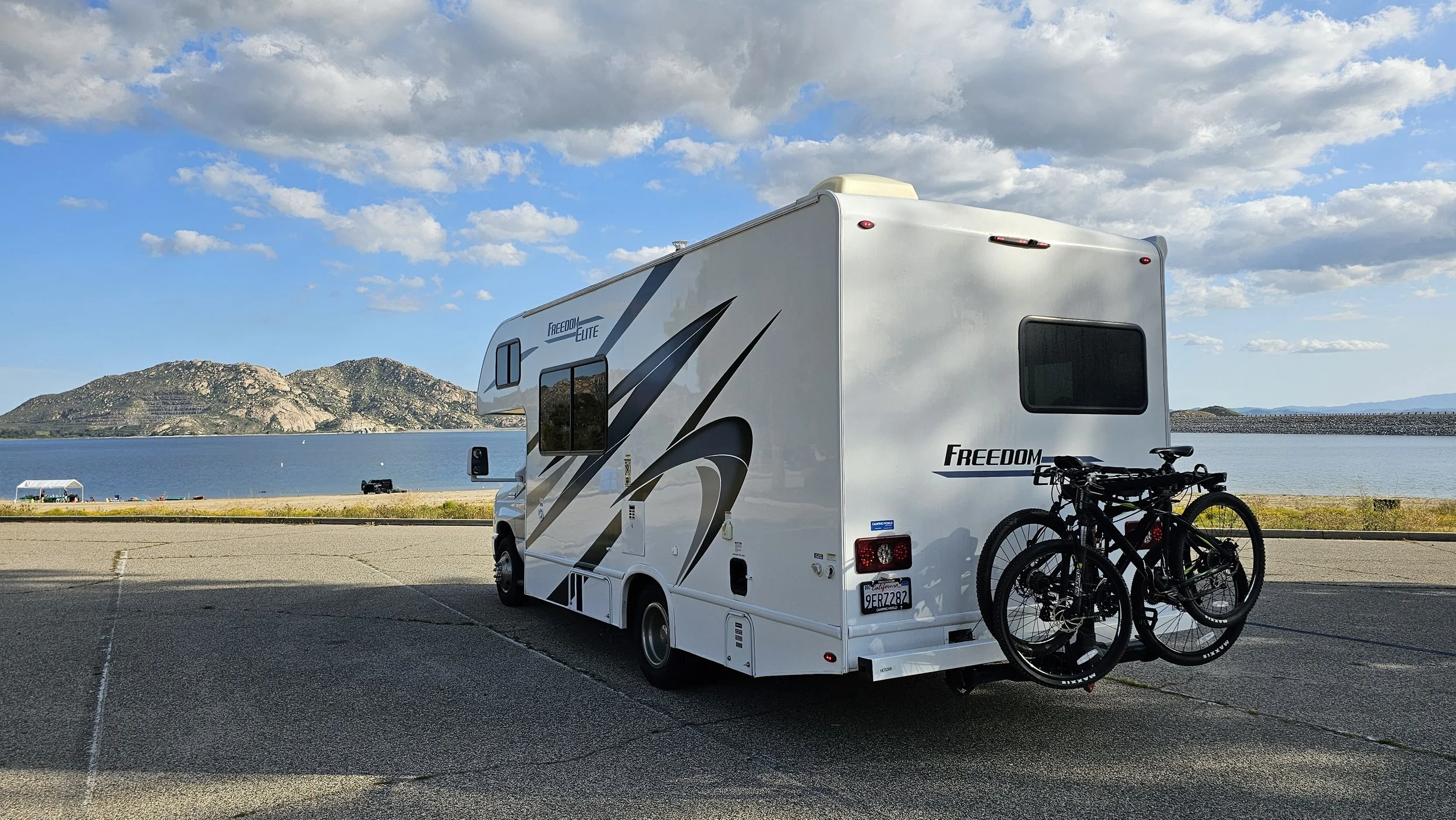 A white recreational vehicle (RV) with two bicycles mounted on the back, parked on a paved lot near a body of water with a mountain range in the background under a partly cloudy sky.