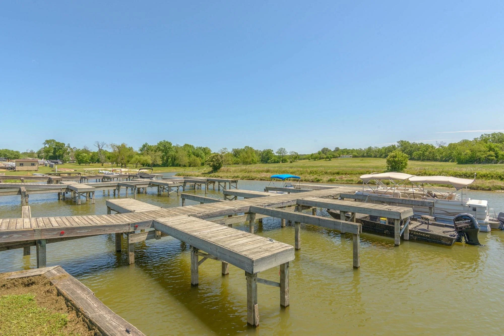 Wooden docks extending over water with small boat and pontoon boat, green trees and blue sky in the background.
